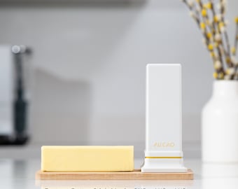 May include: A white butter dish with the text "AU CAO" on the front, next to a stick of butter on a wooden cutting board. The butter stick is 3 1/2 inches by 1 1/2 inches by 1 1/2 inches.
