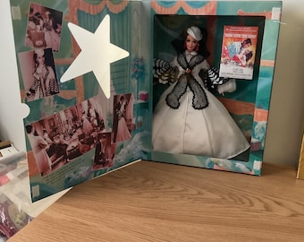 May include: A doll in a box, dressed in a white gown with a black and gray shawl. The box features a star-shaped cutout and images from the movie "Gone With the Wind". The doll has a white hat and is in a clear plastic display.