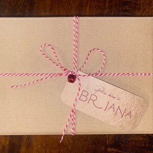 Op de afbeelding: Een kleine, rechthoekige kartonnen doos vastgebonden met rood en wit touw. Een kleine rode bel zit in het midden van de strik. Een bruin label leest "Please deliver to BRIANA" in een cursief lettertype.