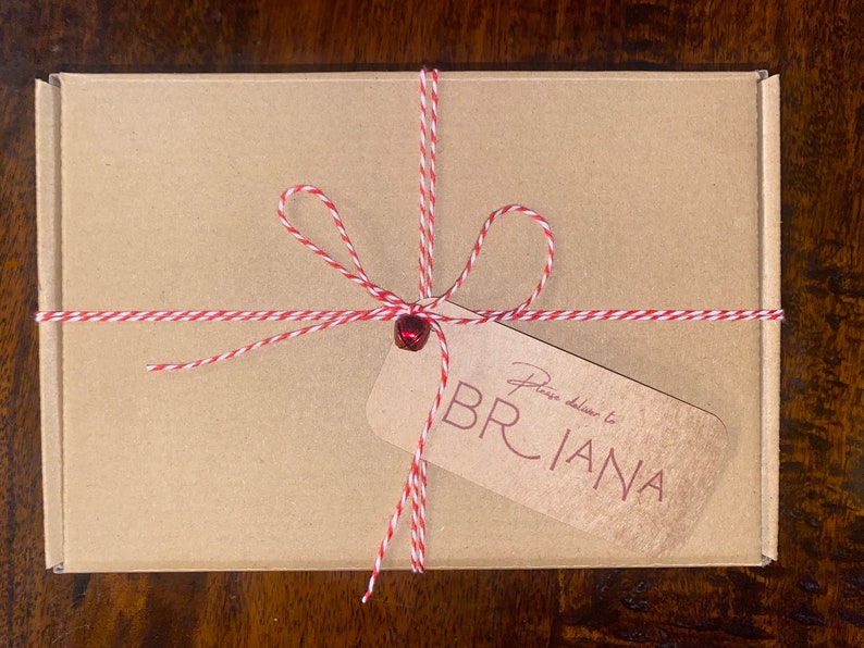 Op de afbeelding: Een kleine, rechthoekige kartonnen doos vastgebonden met rood en wit touw. Een kleine rode bel zit in het midden van de strik. Een bruin label leest "Please deliver to BRIANA" in een cursief lettertype.