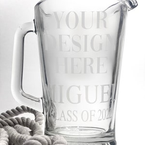 May include: A clear glass pitcher with a handle, etched with the text "YOUR DESIGN HERE MIGUEL CLASS OF 2022". The pitcher is sitting on a gray rope.