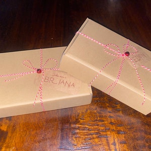 Op de afbeelding: Twee rechthoekige kartonnen dozen, vastgebonden met rood en wit touw en een rode kraal. Op &eacute;&eacute;n doos staat de naam "BRIANA" op een label. Op de andere doos staat de naam "EDEN". De dozen staan op een donker houten oppervlak.