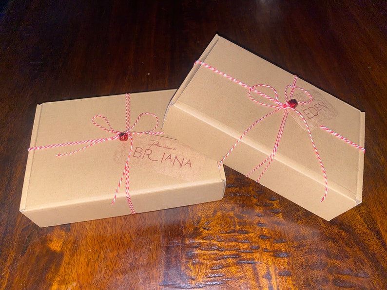 Op de afbeelding: Twee rechthoekige kartonnen dozen, vastgebonden met rood en wit touw en een rode kraal. Op &eacute;&eacute;n doos staat de naam "BRIANA" op een label. Op de andere doos staat de naam "EDEN". De dozen staan op een donker houten oppervlak.