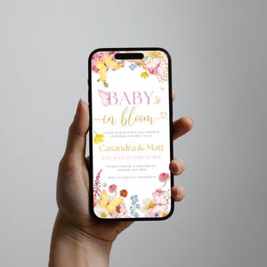 K&ouml;nnte beinhalten: Ein Smartphone zeigt eine Babyparty-Einladung. Das Design zeigt Blumen- und Schmetterlingsillustrationen mit dem Text "Baby in Bloom" in Rosa und Gold. Details f&uuml;r Casandra & Matt sind enthalten.