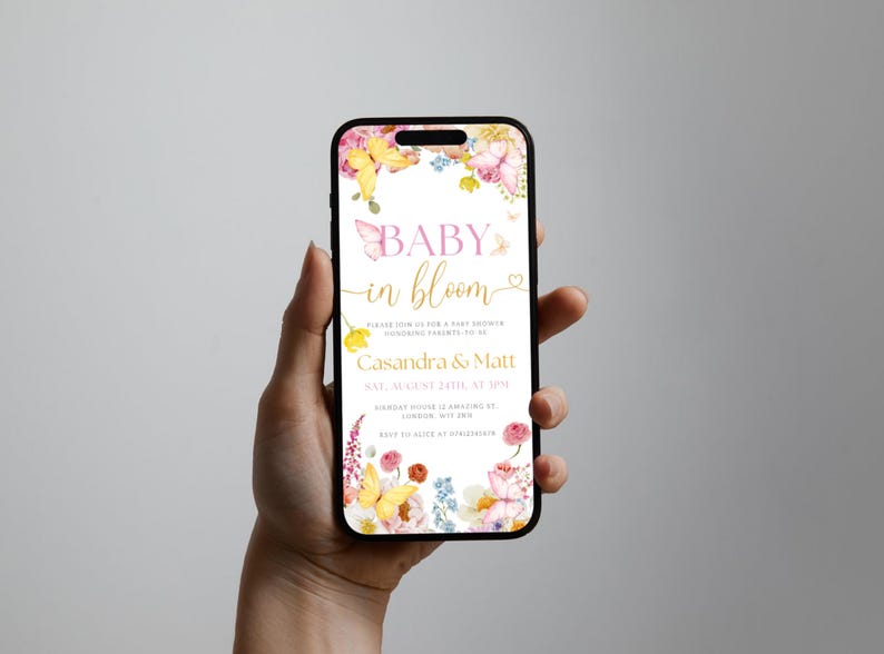 K&ouml;nnte beinhalten: Ein Smartphone zeigt eine Babyparty-Einladung. Das Design zeigt Blumen- und Schmetterlingsillustrationen mit dem Text "Baby in Bloom" in Rosa und Gold. Details f&uuml;r Casandra & Matt sind enthalten.