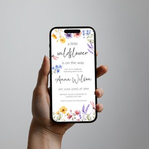 Puede incluir: Una invitaci&oacute;n digital para un baby shower con un borde floral de acuarela. El texto dice "una peque&ntilde;a flor silvestre est&aacute; en camino" y "Anna Wilson". La invitaci&oacute;n incluye la fecha, la hora y la ubicaci&oacute;n del baby shower.