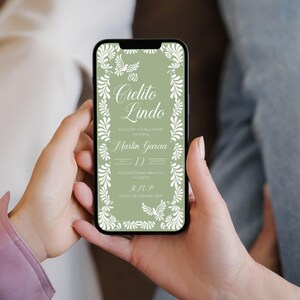 May include: A smartphone displays a wedding invitation with the text "Cielito Lindo" in elegant script, framed by white floral and dove illustrations on a sage green background. The invitation includes details for Martin Garcia's wedding.
