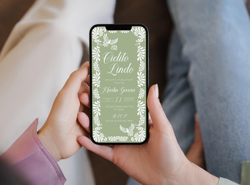 May include: A smartphone displays a wedding invitation with the text "Cielito Lindo" in elegant script, framed by white floral and dove illustrations on a sage green background. The invitation includes details for Martin Garcia's wedding.