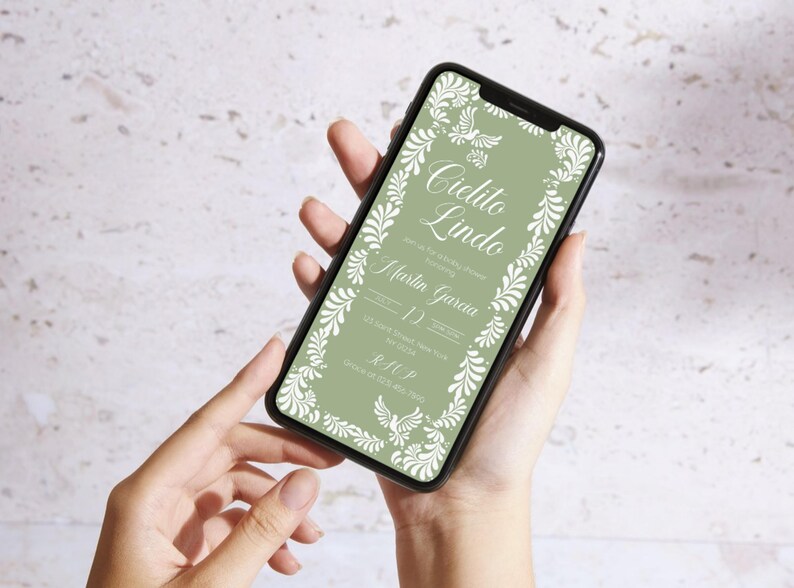 May include: A smartphone displays a digital invitation with the words "Cielito Lindo" in elegant script. The invitation features a sage green background with white floral and bird accents. The invitation includes the names Martin Garcia and the date.