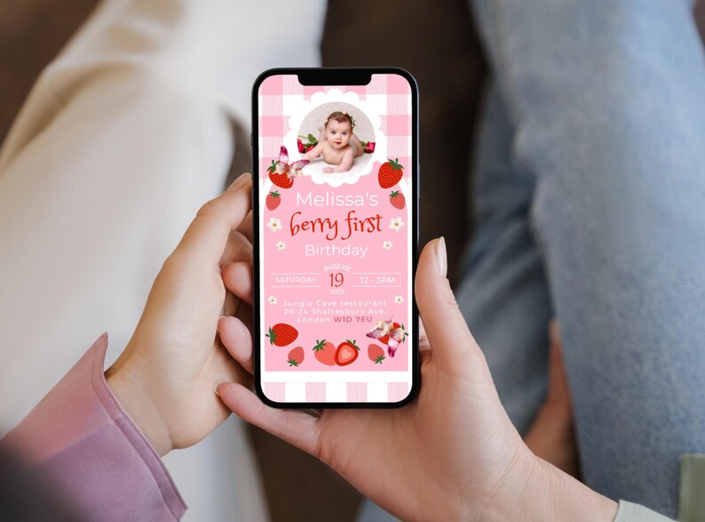 May include: A smartphone displays a pink invitation for "Melissa's berry first Birthday." The design features strawberries, flowers, and a baby's photo. The invitation includes the date, time, and location details, all set against a gingham background.