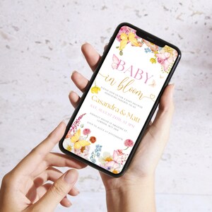 K&ouml;nnte beinhalten: Ein Smartphone zeigt eine Babyparty-Einladung mit Blumen- und Schmetterlingsmotiven. Der Text lautet "Baby in Bloom" mit Details f&uuml;r Casandra & Matts Party am 24. August. Das Telefon wird vor einem hellen Hintergrund gehalten.