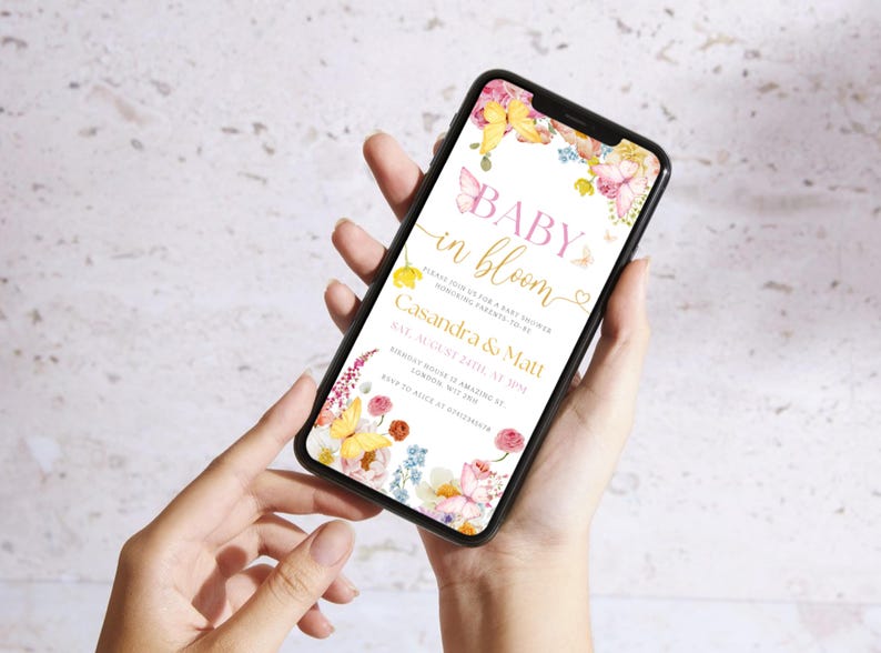 K&ouml;nnte beinhalten: Ein Smartphone zeigt eine Babyparty-Einladung mit Blumen- und Schmetterlingsmotiven. Der Text lautet "Baby in Bloom" mit Details f&uuml;r Casandra & Matts Party am 24. August. Das Telefon wird vor einem hellen Hintergrund gehalten.