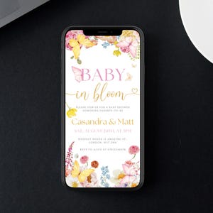 K&ouml;nnte beinhalten: Ein Smartphone zeigt eine Babyparty-Einladung. Der Text "BABY in bloom" ist in Rosa und Gold gehalten und von Aquarellblumen und Schmetterlingen umgeben. Die Einladung enth&auml;lt die Namen der Eltern, Datum, Uhrzeit und Ort.