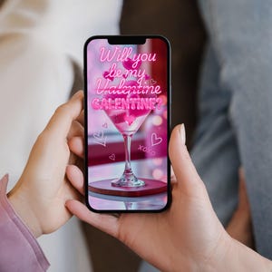 Op de afbeelding: Een smartphone toont een Valentijnsdag-afbeelding. De afbeelding toont een roze cocktail in een martiniglas, omringd door harten en de tekst "Will you be my Valentine/Galentine?" in een neon-stijl lettertype. De telefoon wordt door een persoon vastgehouden.