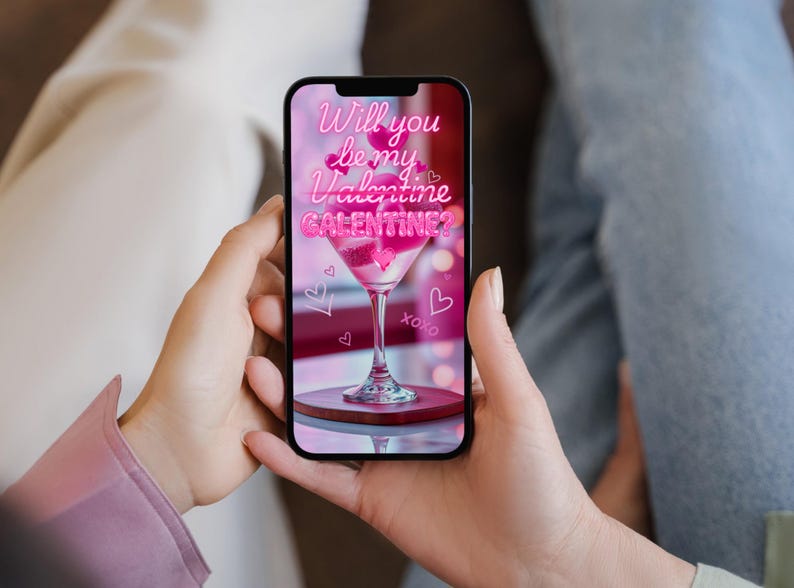 Op de afbeelding: Een smartphone toont een Valentijnsdag-afbeelding. De afbeelding toont een roze cocktail in een martiniglas, omringd door harten en de tekst "Will you be my Valentine/Galentine?" in een neon-stijl lettertype. De telefoon wordt door een persoon vastgehouden.