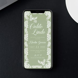 May include: A smartphone displays a baby shower invitation with the text "Cielito Lindo" in elegant script. The invitation features a sage green background with white floral and dove accents. The phone is on a black surface.