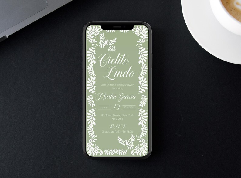 May include: A smartphone displays a baby shower invitation with the text "Cielito Lindo" in elegant script. The invitation features a sage green background with white floral and dove accents. The phone is on a black surface.