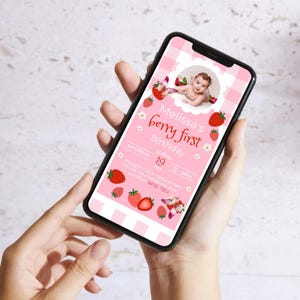 May include: A smartphone displays a pink digital invitation for a first birthday party. The invitation features a baby's photo, strawberries, and the text "Melissa's berry first Birthday". The phone is held in a person's hands.