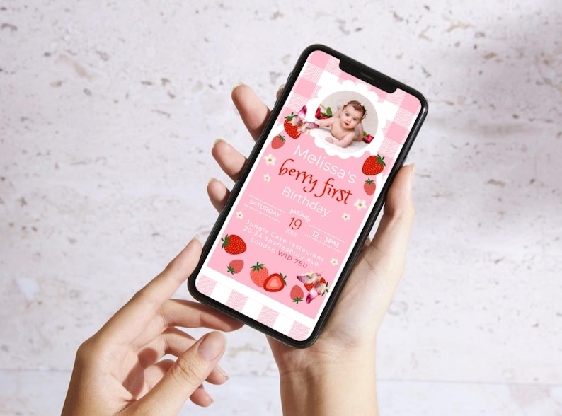 May include: A smartphone displays a pink digital invitation for a first birthday party. The invitation features a baby's photo, strawberries, and the text "Melissa's berry first Birthday". The phone is held in a person's hands.