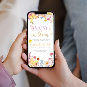 K&ouml;nnte beinhalten: Ein Smartphone zeigt eine Babyparty-Einladung mit Blumen- und Schmetterlingsmotiven. Der Text lautet "Baby in Bloom" mit Details f&uuml;r Cassandra & Matt, am 24. August um 13:00 Uhr.