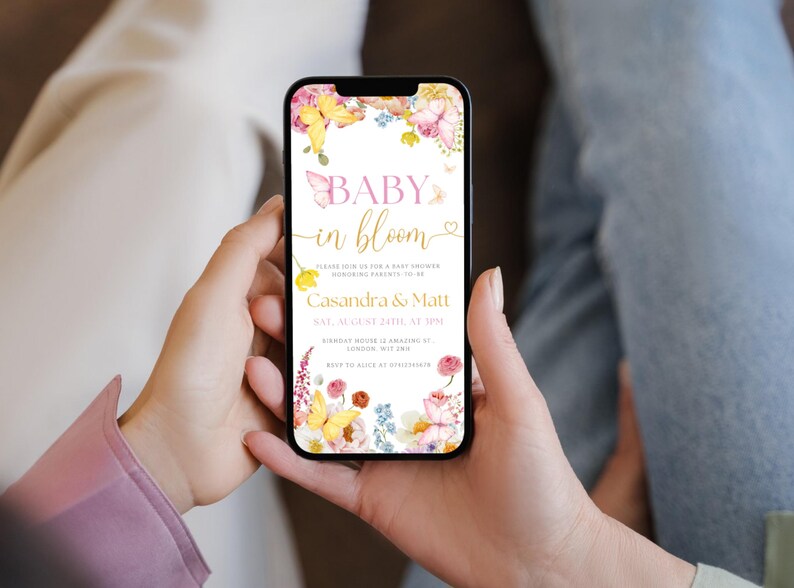 K&ouml;nnte beinhalten: Ein Smartphone zeigt eine Babyparty-Einladung mit Blumen- und Schmetterlingsmotiven. Der Text lautet "Baby in Bloom" mit Details f&uuml;r Cassandra & Matt, am 24. August um 13:00 Uhr.