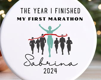May include: A round white ceramic ornament featuring the black text THE YEAR I FINISHED MY FIRST MARATHON, a graphic of runners in black and a central runner in light green crossing a red finish line, with the name Sabrina and the year 2024 below.