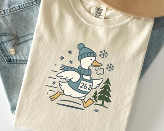 May include: A cream-colored t-shirt featuring a graphic of a white duck wearing a blue beanie, scarf, and a race bib with the number 26.2. The duck is running past a green pine tree amidst falling snowflakes, paired with light blue denim jeans.