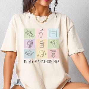 May include: A beige short-sleeve t-shirt featuring a grid of nine pastel-colored squares, each containing a black line-art icon related to running, including a shoe, water bottle, watch, and medal, with the text IN MY MARATHON ERA printed below.