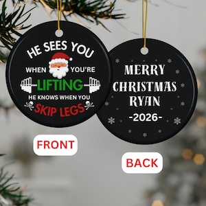 May include: Two round black ceramic ornaments with gold hanging strings. The front features a Santa graphic with text: HE SEES YOU WHEN YOU'RE LIFTING HE KNOWS WHEN YOU SKIP LEGS. The back displays MERRY CHRISTMAS RYAN -2026- with white snowflake icons.