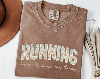 May include: A brown Comfort Colors t-shirt with the word "RUNNING" in a floral design. Below the word is the phrase "because it's cheaper than therapy". A silver necklace with a small pendant is on the shirt.