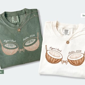 May include: Two t-shirts, one moss green and one ivory, with a coconut bra graphic. The graphic includes the text "Megan's Back Malibu, 2026". Each shirt has a gold necklace. The shirts are from Comfort Colors.
