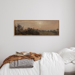 May include: A framed landscape painting depicting a rural scene with trees, hills, and a body of water in the distance. The sky is a soft pink and orange, suggesting a sunset or sunrise. The painting is in a wooden frame with a light brown finish.