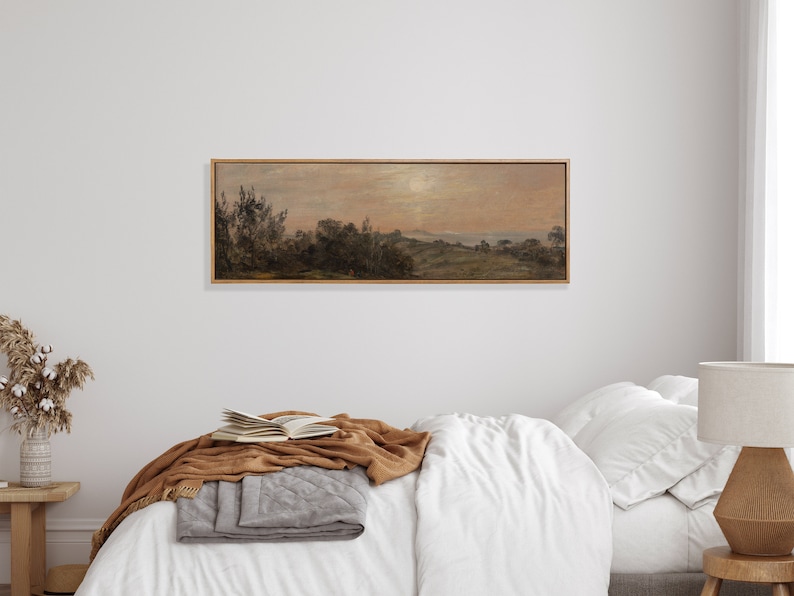May include: A framed landscape painting depicting a rural scene with trees, hills, and a body of water in the distance. The sky is a soft pink and orange, suggesting a sunset or sunrise. The painting is in a wooden frame with a light brown finish.
