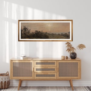May include: A framed landscape painting depicting a rural scene with trees, hills, and a body of water in the distance. The painting is in a light brown wooden frame and is hanging on a white wall above a wooden cabinet.
