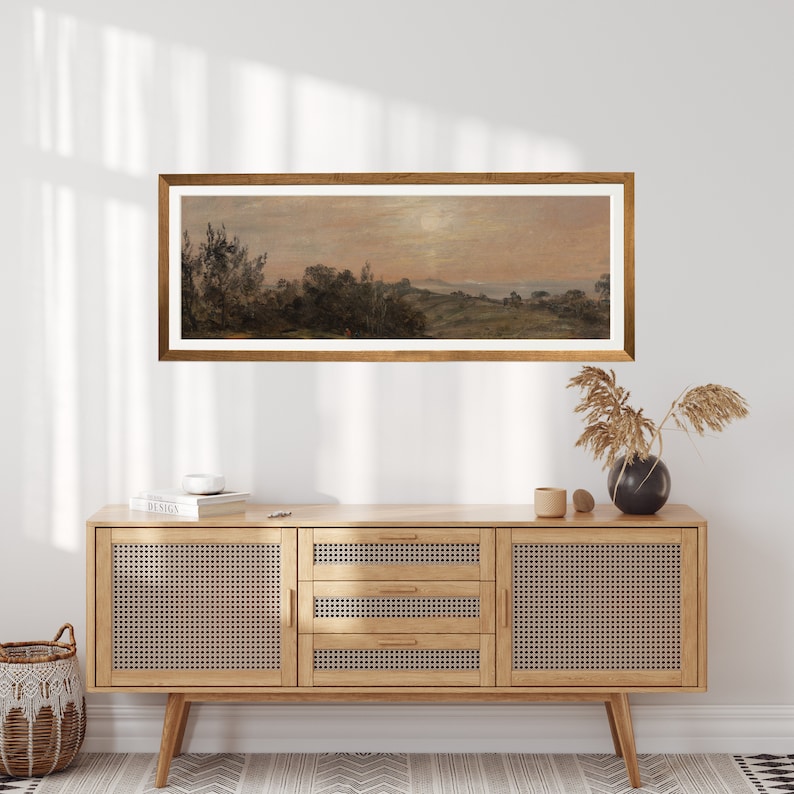 May include: A framed landscape painting depicting a rural scene with trees, hills, and a body of water in the distance. The painting is in a light brown wooden frame and is hanging on a white wall above a wooden cabinet.