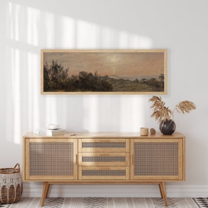 May include: A framed landscape painting of a rural scene with trees, hills, and a body of water in the distance. The painting is in a light wood frame and is hanging on a white wall above a wooden cabinet with woven cane doors.