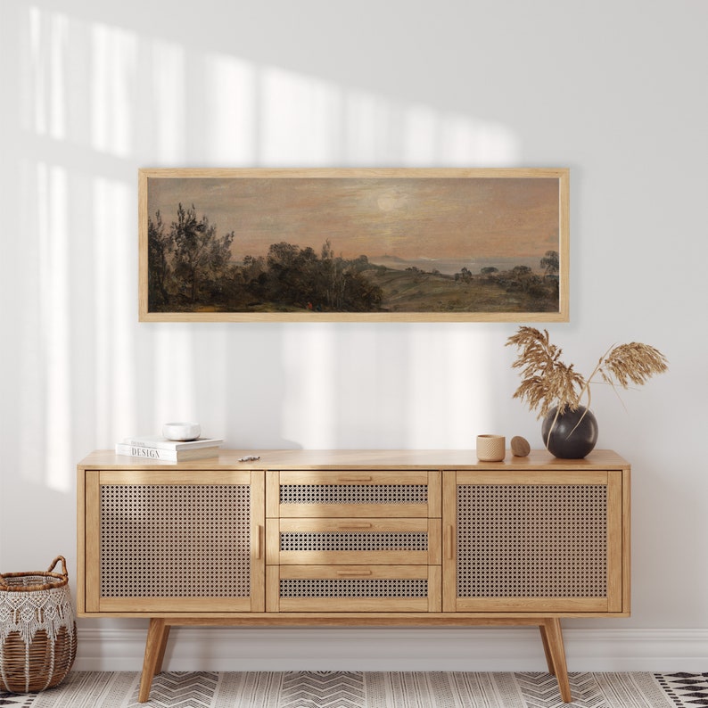 May include: A framed landscape painting of a rural scene with trees, hills, and a body of water in the distance. The painting is in a light wood frame and is hanging on a white wall above a wooden cabinet with woven cane doors.