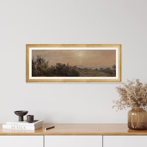 May include: A framed landscape painting depicting a rural scene with trees, hills, and a body of water in the distance. The sky is a soft pink and orange, suggesting a sunset or sunrise. The painting is in a gold frame.