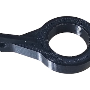 Puede incluir: Una pieza negra, impresa en 3D, con un anillo circular y un pequeño brazo rectangular con un agujero. La pieza está hecha de un material sólido y mate, y parece ser un componente mecánico o conector.
