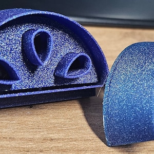 Puede incluir: Cortador de galletas azul de dos piezas, semicircular, con una textura brillante. Una pieza tiene tres recortes en forma de lágrima. La otra pieza es un semicírculo a juego. El cortador es de plástico.