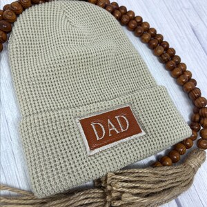 May include: A beige knitted beanie with a brown leather patch that says "DAD" on it. The beanie has a brown tassel.