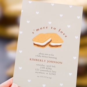 May include: A baby shower invitation with a light beige background and white heart accents. The invitation features the text "s'more to love" above an illustration of two s'mores. Details include the honoree's name, date, time, and location.