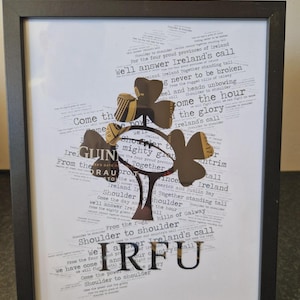 May include: A framed print with a black frame and a white background. The print features a gold shamrock with the words "Come the hour, come the glory, Ireland's call" and "Shoulder to shoulder" repeated throughout the design.