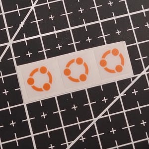 Könnte beinhalten: Drei quadratische Aufkleber in einer horizontalen Reihe, jeder mit einem orangefarbenen kreisförmigen Logo, das aus drei verbundenen Punkten besteht, die einen Ring bilden. Die Aufkleber befinden sich auf einer transparenten Trägerfolie.