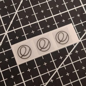 Op de afbeelding: Een strook met drie kleine, vierkante, doorzichtige stickers, elk voorzien van een zwarte cirkelvormige omtrek met een gestileerde kleine letter e. De stickers zijn horizontaal gerangschikt op een witte achtergrondstrook.