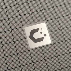 May include: A square, silver sticker with a dark grey geometric design. The design features a stylised letter "C" shape with three small circles to the right. The sticker is on a grey grid background.