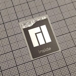 Könnte beinhalten: Ein rechteckiger schwarzer Aufkleber mit weißem Logo und dem Wort "inside" in Weiß gedruckt. Der Aufkleber hat einen silbernen Rand und befindet sich auf einem grauen Rasterhintergrund.
