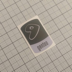 May include: A sticker with a dark gray background and a light purple bottom section. The sticker features a white heart-shaped design with a small circle inside. The word "gentoo" is printed in gray.