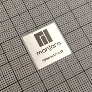 May include: A square sticker with the word "manjaro" in a sans-serif font, and the words "open source" below it. The sticker has a gray and black logo at the top. The sticker is on a gray grid background.