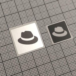 Könnte beinhalten: Zwei quadratische Aufkleber mit einem Fedora-Hut-Design. Einer hat einen silbernen Hintergrund mit einem dunkelgrauen Hut und einem weißen Band. Der andere hat einen schwarzen Hintergrund mit einem weißen Hut.
